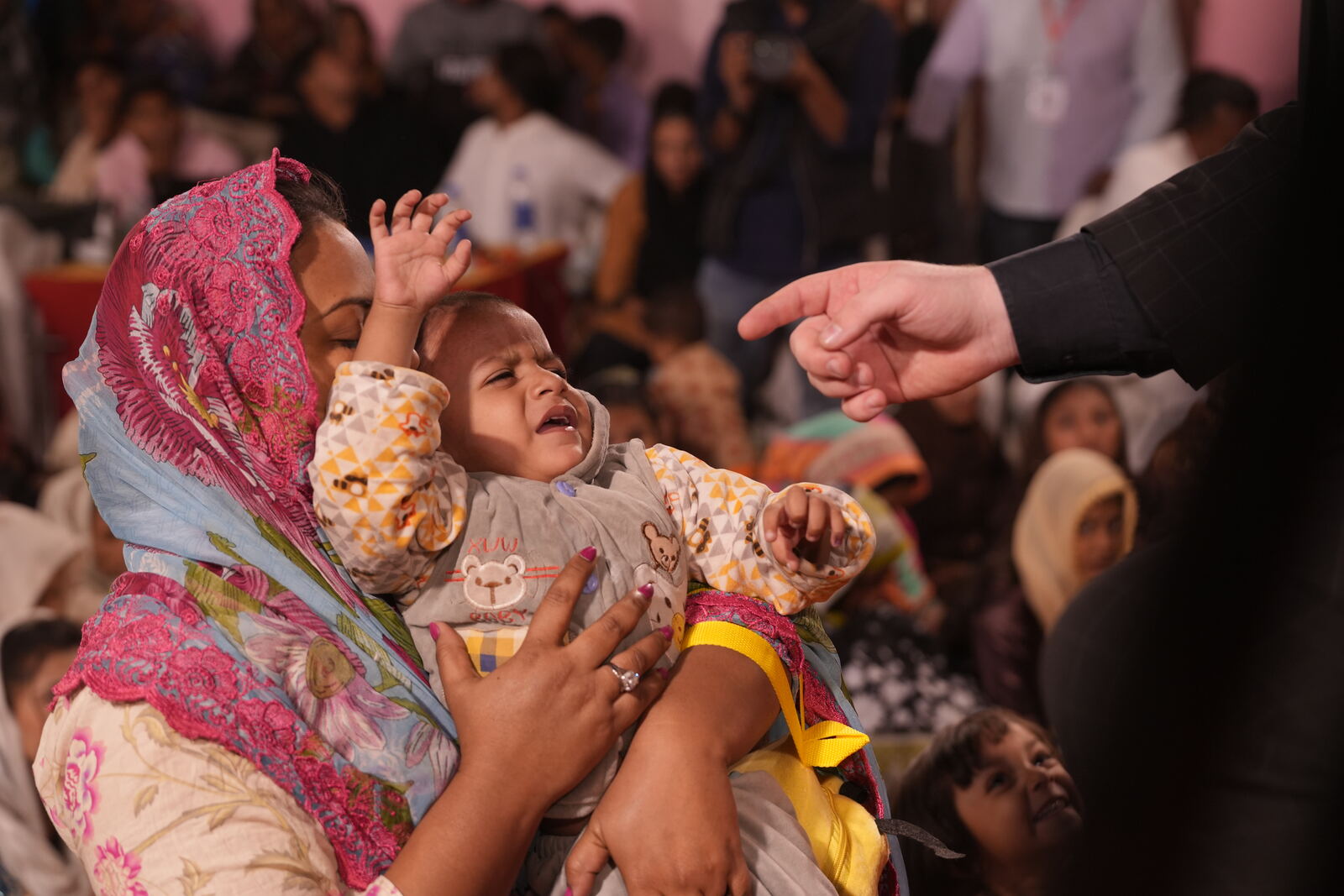 A screaming baby held by his mother as hands reach out to pray for him